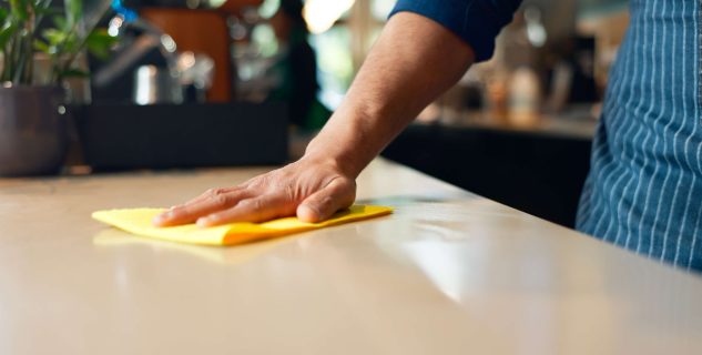 Waiter, hands and cleaning table in coffee shop for dust, bacteria and dirt with cloth, spray or detergent. Barista, person and wipe wooden furniture in cafe or restaurant for shine or professional.